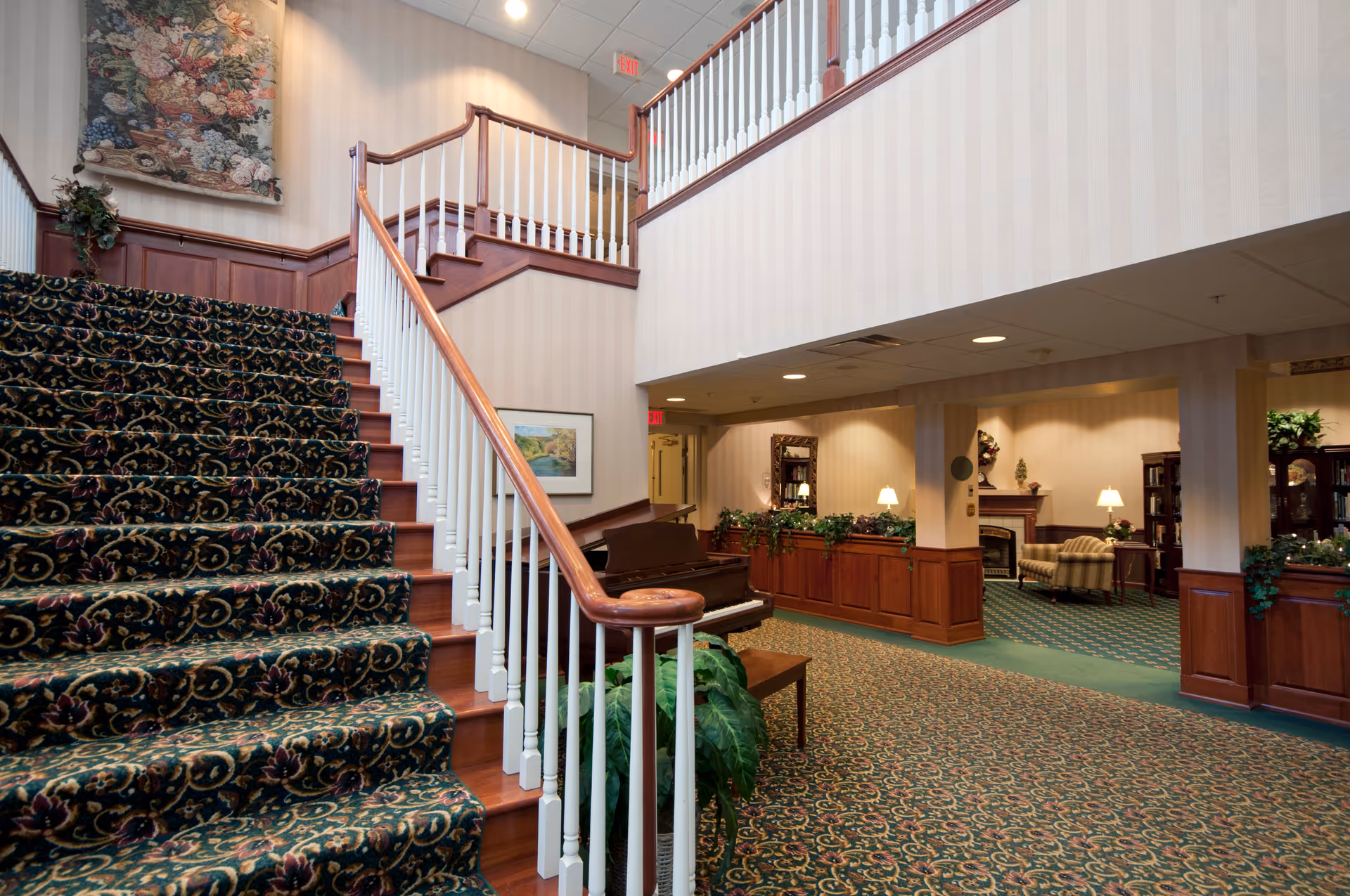 Interior view of a senior living facility featuring a carpeted staircase with a wooden handrail and white balusters. The area includes a piano, green plants, and a seating area with armchairs, lamps, and a fireplace. The walls have light-colored wallpaper and wood paneling, with framed artwork and decorative elements.