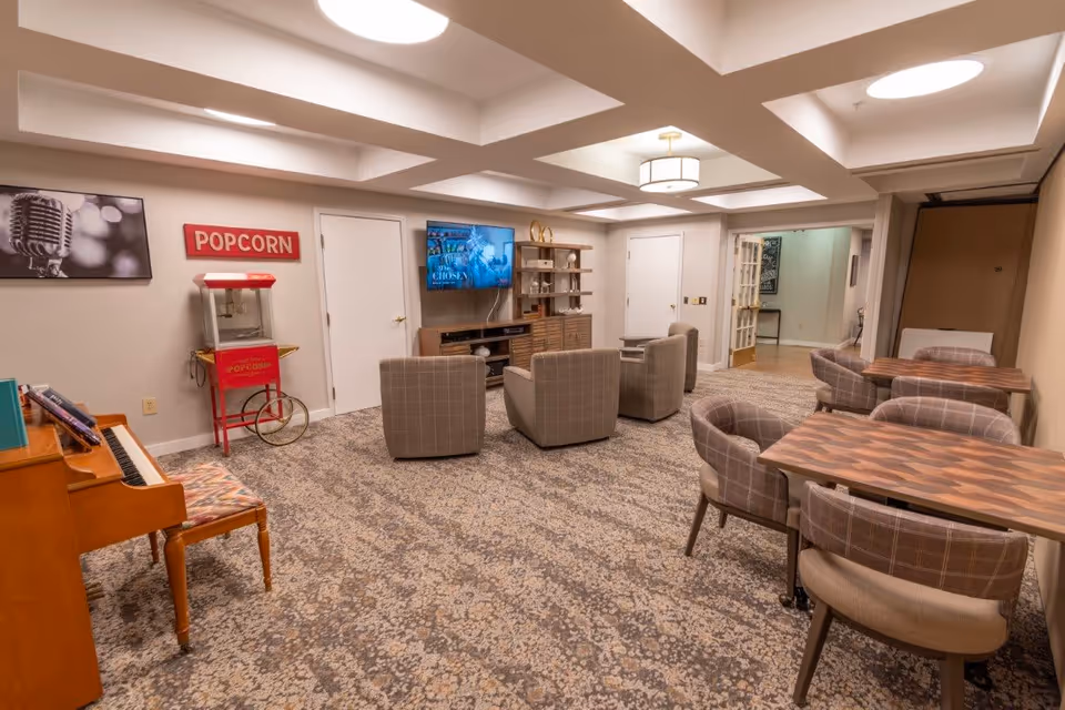 A cozy common area in a senior living facility featuring a popcorn machine, a piano with a bench, several cushioned armchairs arranged around a TV mounted on the wall, and two wooden tables with upholstered chairs. The room has patterned carpet, a ceiling with recessed lighting, and neutral-colored walls.