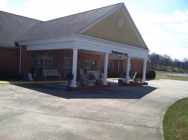 Front exterior view of Maplewood Lane Assisted Living facility with a covered entrance supported by white columns, benches on the porch, and a paved driveway in front.