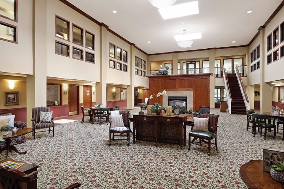 Spacious senior living facility common area with patterned carpet, multiple seating arrangements including chairs and tables, a central wooden table with decorative items, a large fireplace with wooden paneling, and a staircase leading to an upper level with windows and railings.