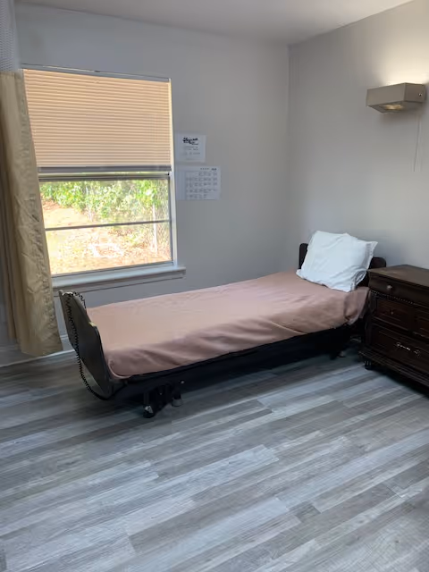 A simple bedroom with a single bed covered with a light pink sheet and a white pillow. Next to the bed is a dark wooden nightstand with drawers. A window with closed blinds and a curtain is on the left wall, letting in some natural light. The floor has gray wood-like vinyl planks, and the walls are painted light gray. A wall-mounted light fixture is above the nightstand.