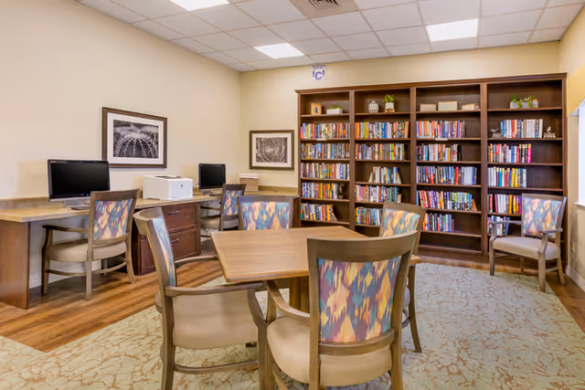 A community library room with a wooden table and chairs, bookshelves filled with books, and computer stations along the wall.