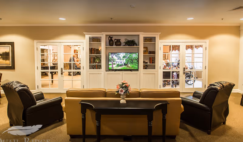 A cozy living room area in an assisted living facility featuring a tan sofa with a small black table behind it holding a vase of flowers, flanked by two black leather armchairs. In front of the seating area is a built-in white entertainment center with shelves holding books and decorative items, and a flat-screen TV displaying an outdoor scene. Through glass double doors on either side of the entertainment center, people in wheelchairs are visible in adjacent rooms.