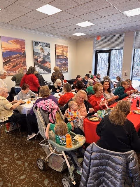 A large group of elderly people and a few children gathered in a bright room with large windows, sitting at tables covered with white and red tablecloths, eating and socializing. The room has posters on the wall featuring Michigan scenes and a carpeted floor.