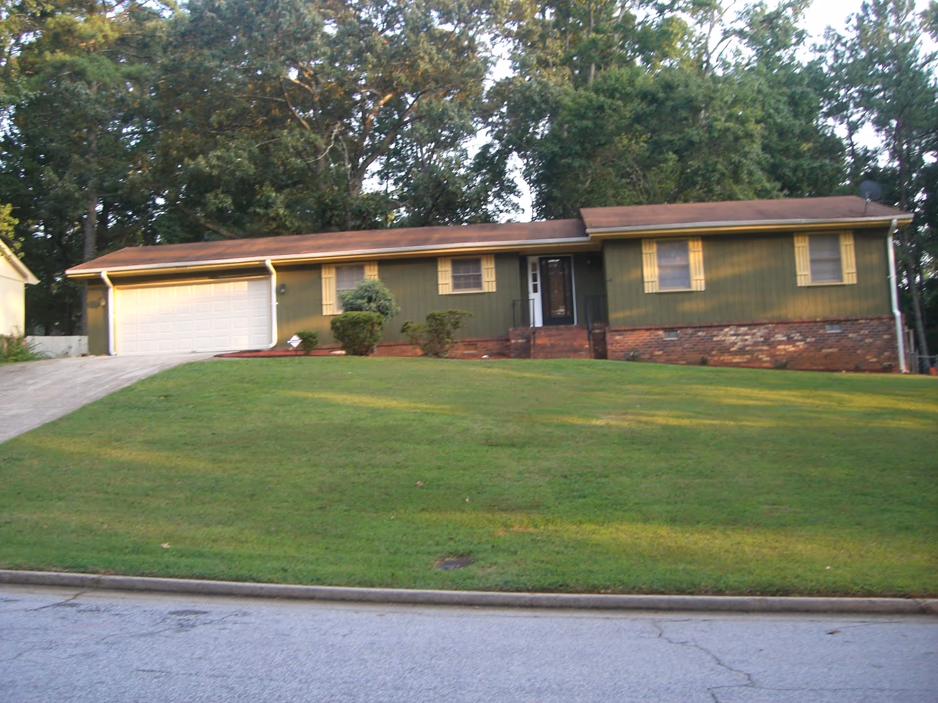 Single-story residential building with green exterior walls, yellow window shutters, a brown roof, and a white garage door. The house is situated on a grassy lawn with a driveway leading to the garage. Trees are visible in the background.