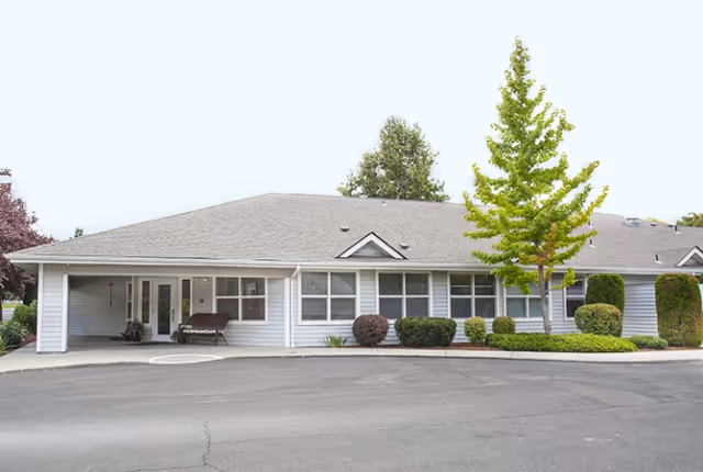 Front exterior of a single-story assisted living building with a covered entrance, bench, large windows, shrubs, and a paved driveway.