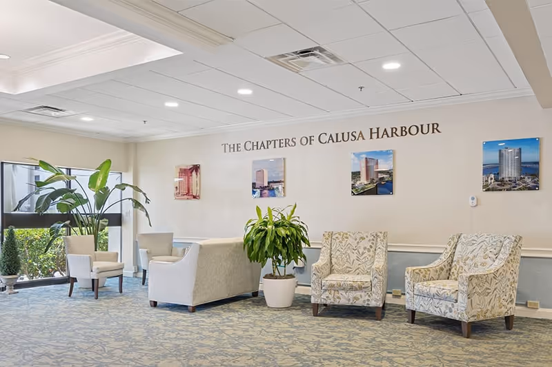 A bright and spacious seating area in a senior living facility with several upholstered armchairs and a sofa arranged around a potted plant. Large windows let in natural light, and framed photos of buildings are displayed on the wall under the text 'The Chapters of Calusa Harbour'.