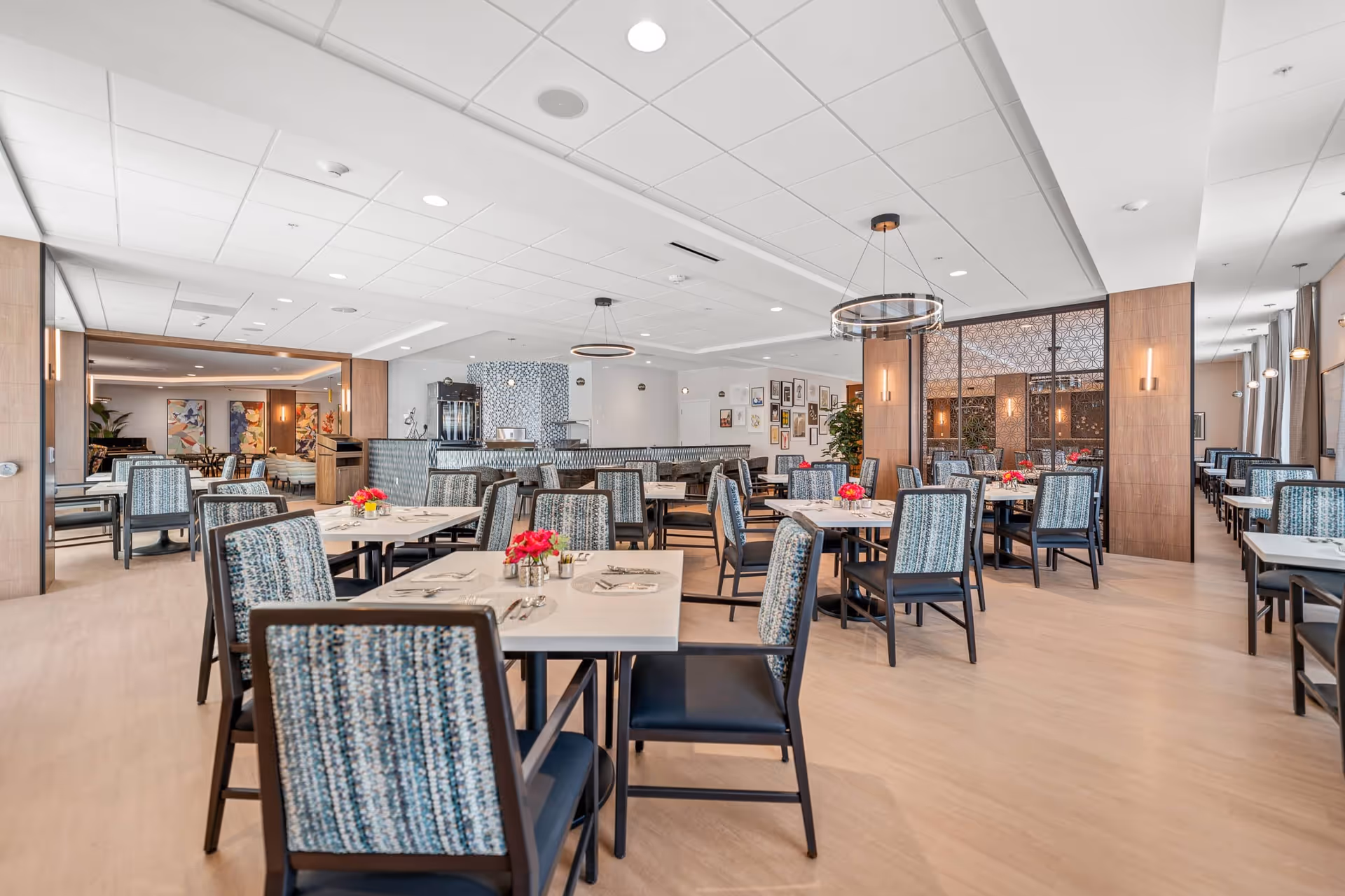 A spacious, well-lit dining room with multiple tables and chairs arranged neatly. Each table is set with placemats, utensils, and small flower arrangements. The room features modern lighting fixtures, light wood flooring, and a decorative partition wall. The overall atmosphere is clean and inviting.