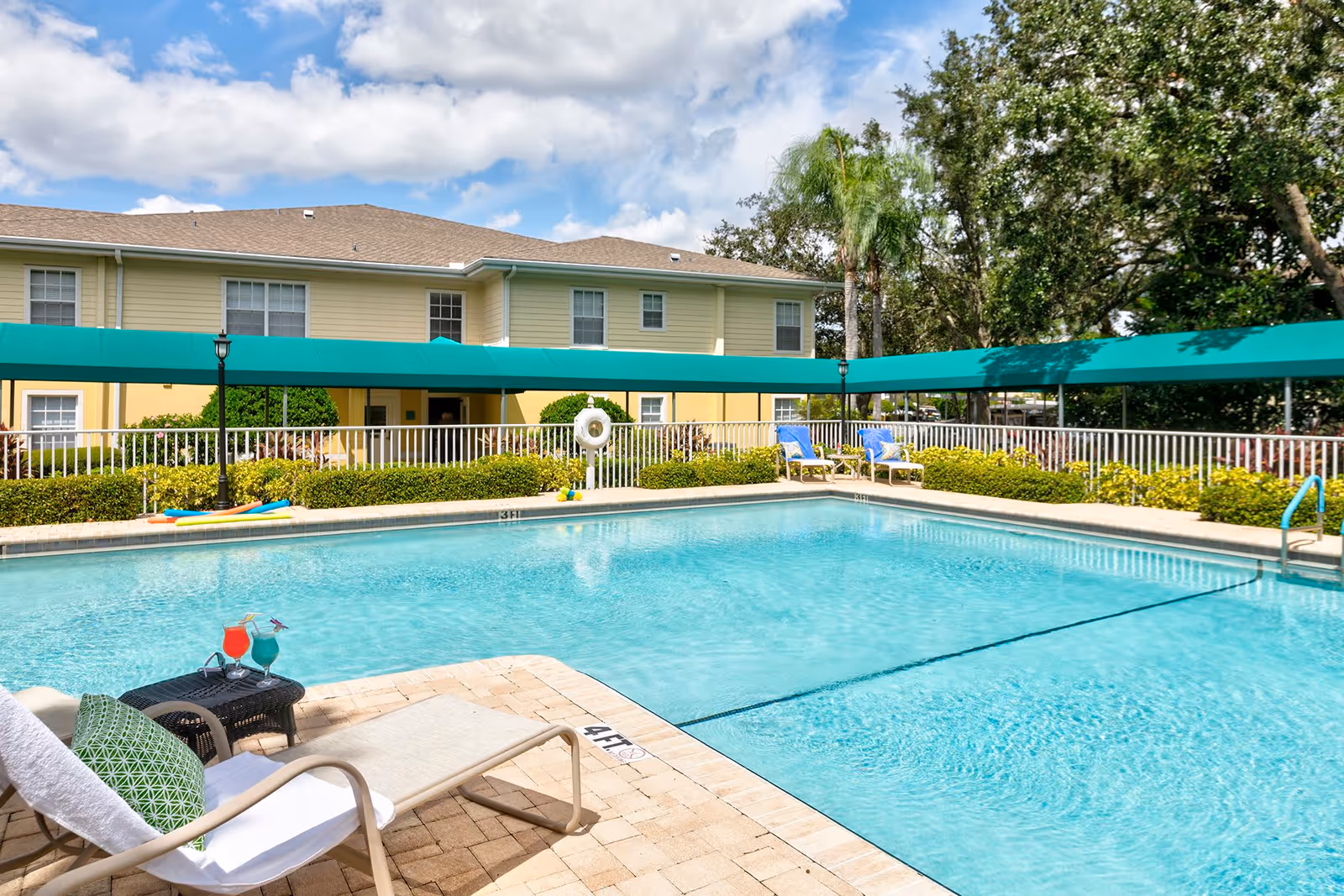 Outdoor swimming pool with clear blue water surrounded by a paved deck. There are lounge chairs with cushions and two colorful drinks on a small table near the pool. In the background, there is a two-story yellow building with white trim and a green awning. Trees and bushes surround the pool area under a partly cloudy sky.