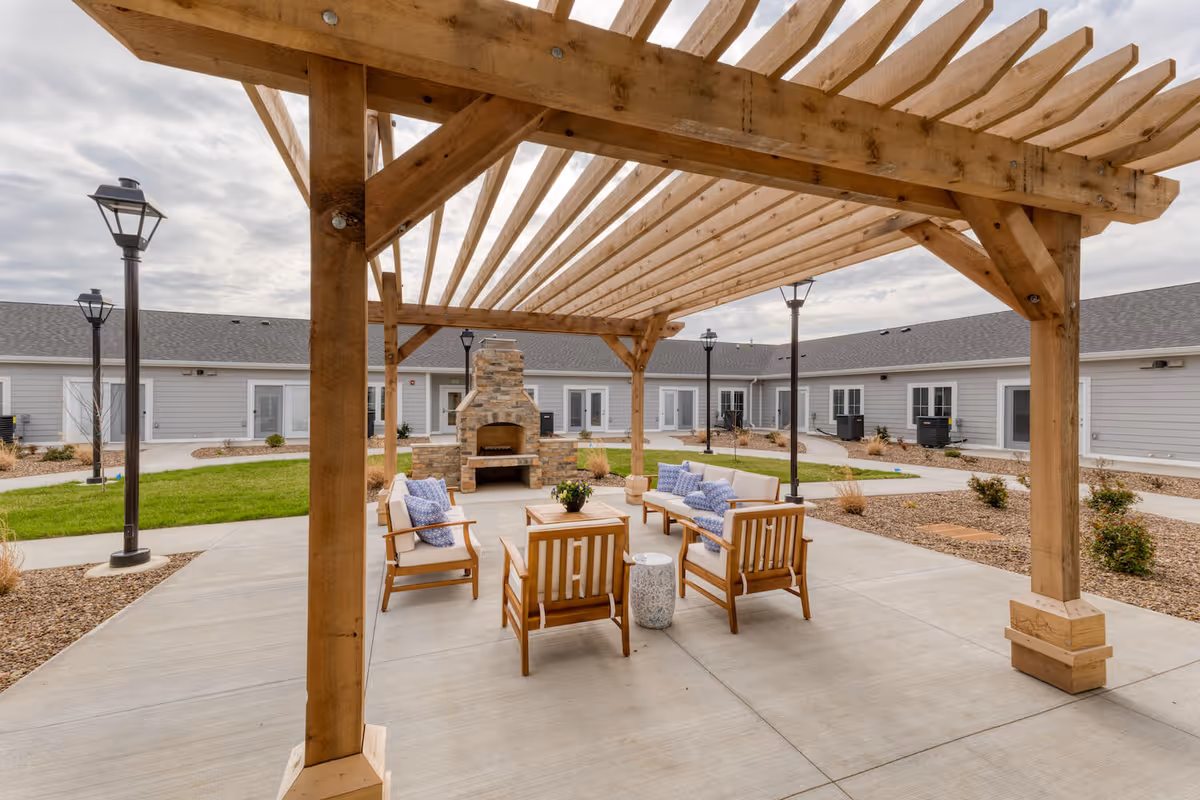 Outdoor seating area at Cedarhurst Senior Living of Owensboro featuring wooden pergola, cushioned wooden chairs and sofas with blue patterned pillows, a stone fireplace, and surrounding pathways with grass and landscaping.
