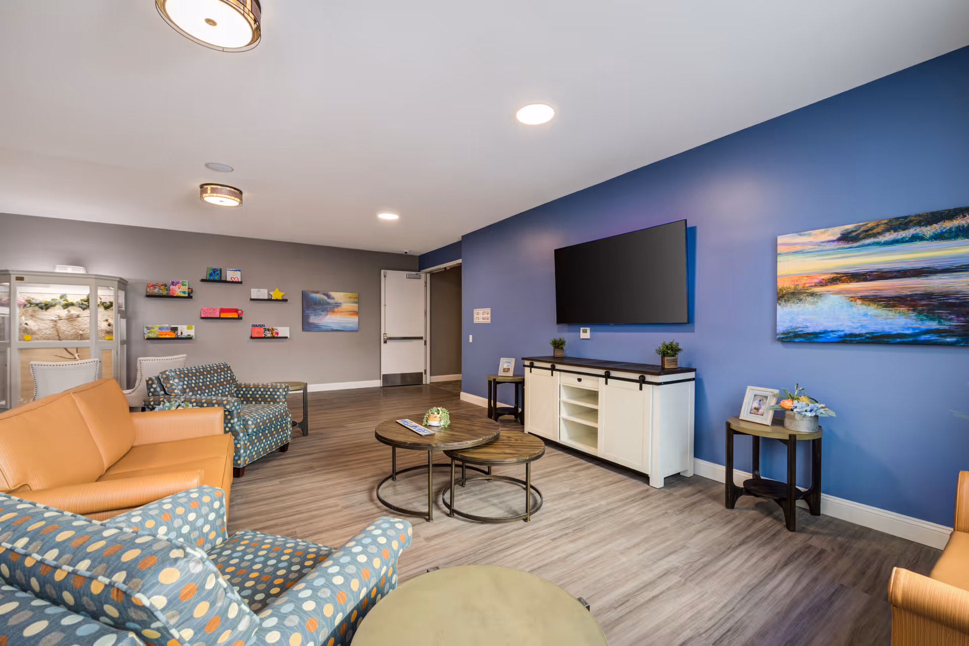 Bright communal living room with sofas, patterned armchairs, a wall-mounted TV, coffee tables, and colorful artwork.