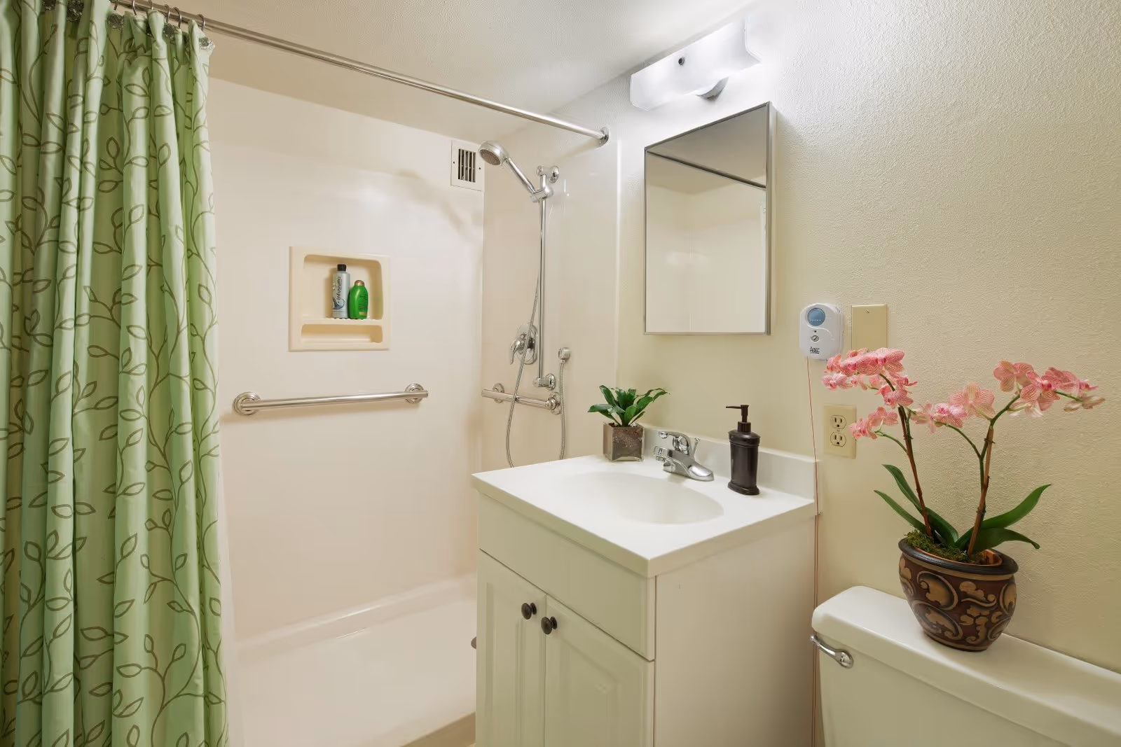 A clean bathroom with a white vanity and sink, a mirror above the sink, a soap dispenser, and a small potted plant. To the right is a toilet with a decorative pot containing pink orchids on top. The shower area has a green shower curtain with a leaf pattern, a handheld showerhead, and grab bars for safety. There are two bottles of shower products in a built-in shower niche.