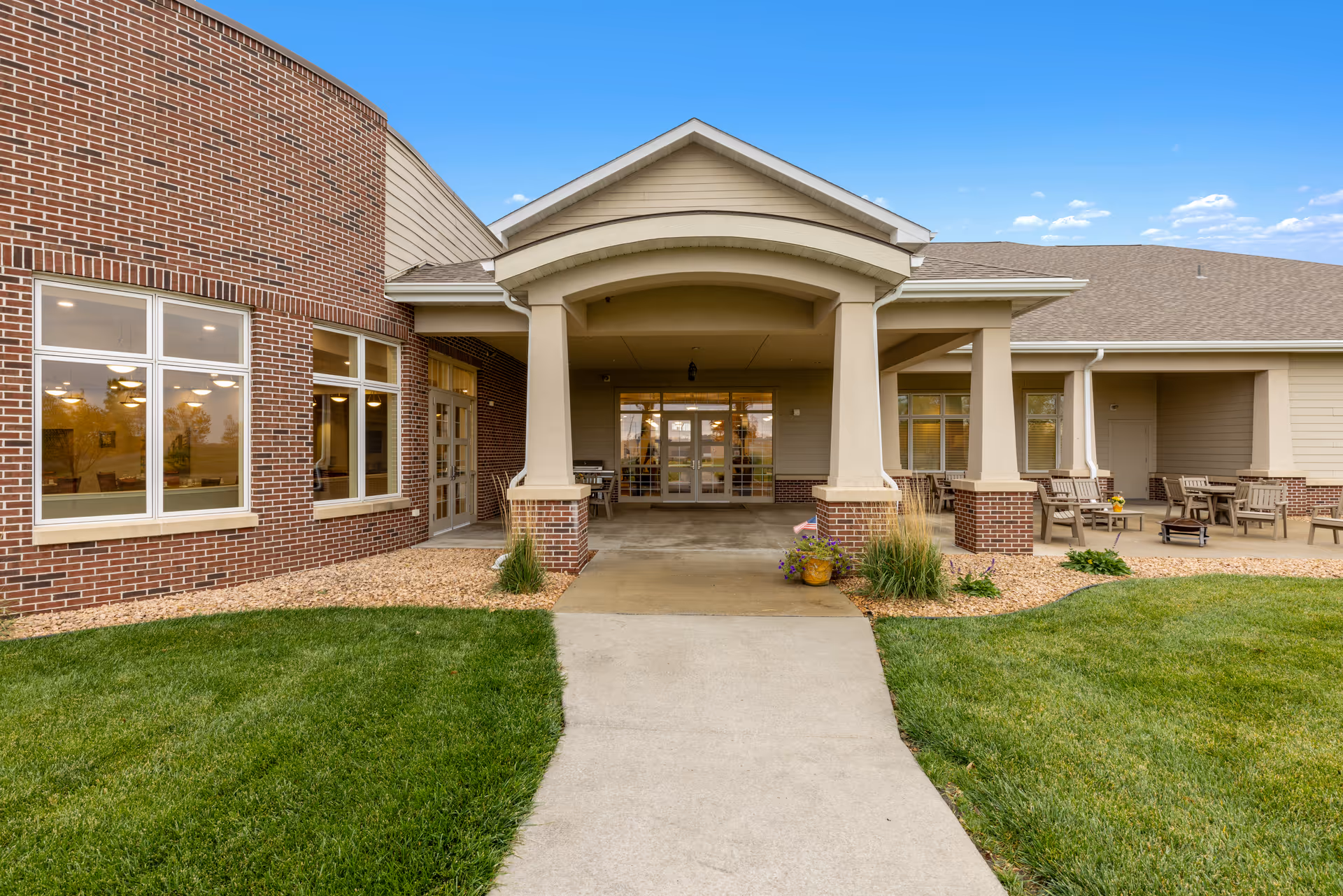 Front exterior view of a senior living facility with a covered entrance supported by columns, brick and beige siding walls, large windows, and outdoor seating areas with chairs and tables on a concrete patio surrounded by green grass and landscaping.
