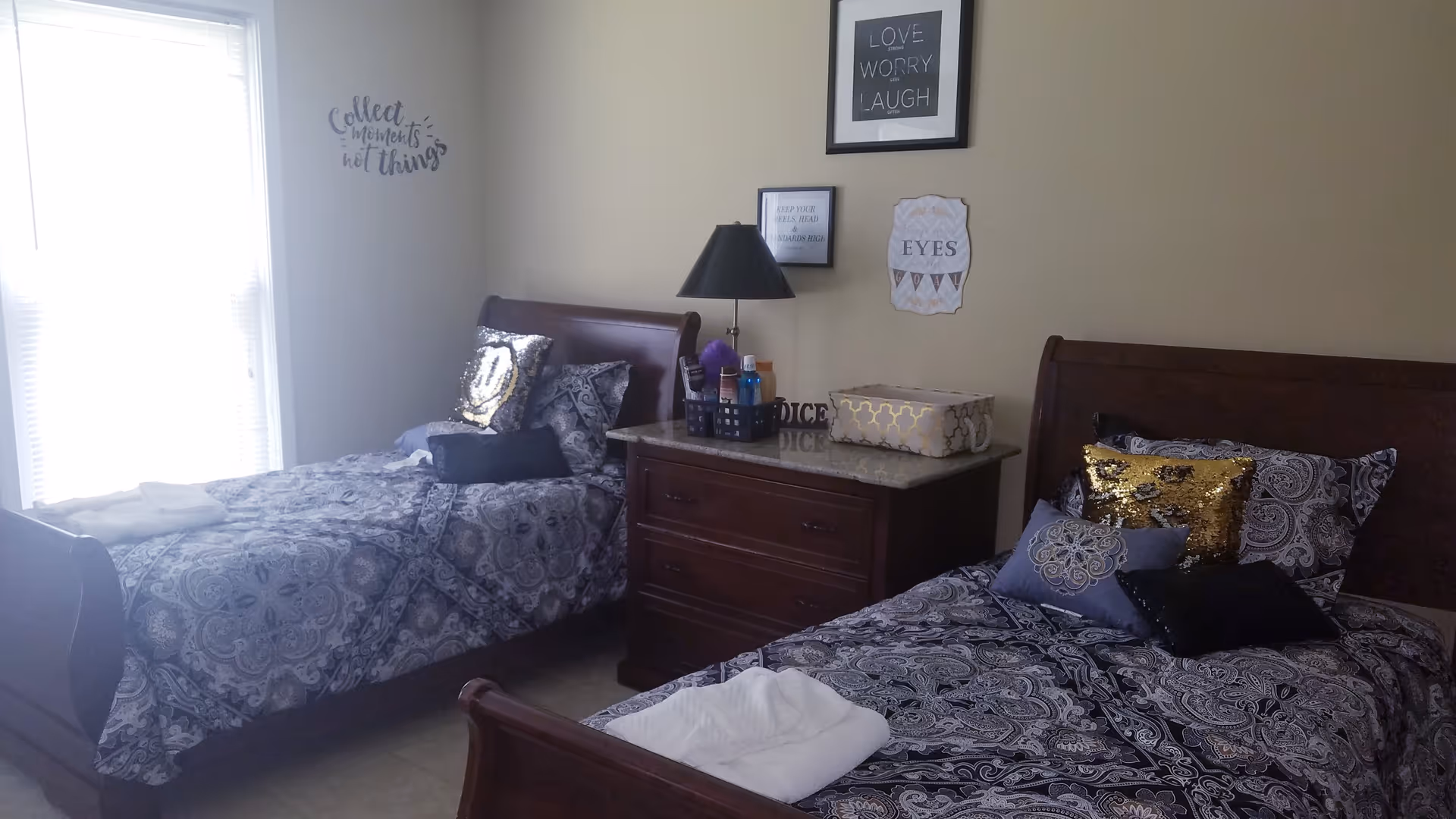 A bedroom with two twin beds featuring dark wooden frames and patterned bedding. Each bed has multiple decorative pillows, including shiny gold and black ones. Between the beds is a dark wooden nightstand with a lamp and various items on top. The walls are light-colored and decorated with framed inspirational quotes and wall art. A window with blinds is on the left side, letting in natural light.