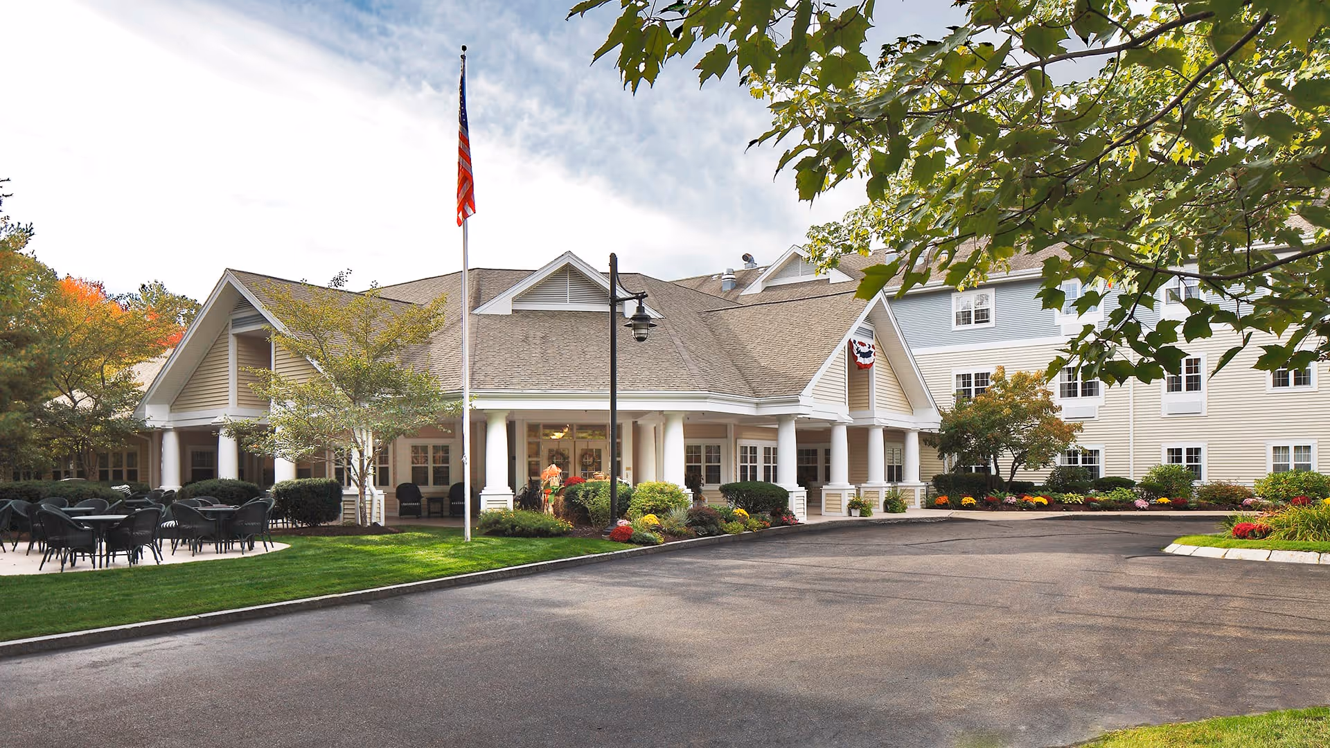 Exterior view of a senior living facility with a large driveway, manicured lawn, outdoor seating area with tables and chairs, an American flag on a flagpole, and a building with white columns and beige siding surrounded by trees and shrubs.