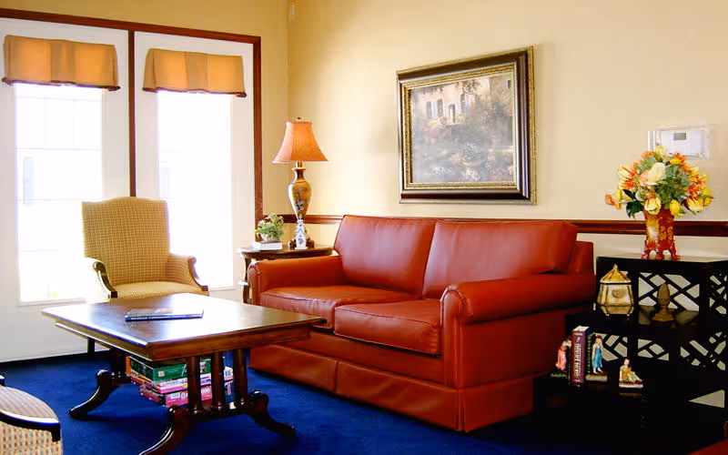 A cozy living room with a red leather sofa, a wooden coffee table with board games underneath, a beige armchair, a side table with a lamp and small plant, a framed painting on the wall, and a decorative shelf with flowers and figurines.