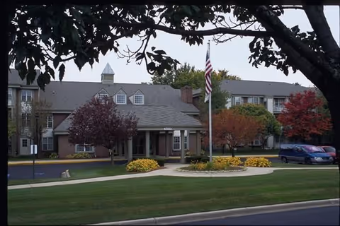 Front entrance of a multi-story senior living building with a flagpole, circular landscaped driveway, and autumn trees.