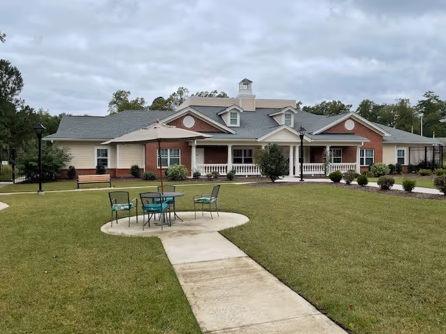 Front exterior of a single-story brick and siding senior living building with a grassy lawn, walkway, and outdoor table and chairs.