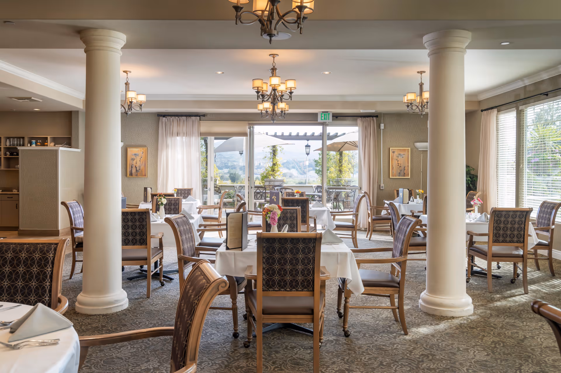 A bright and elegant dining room with multiple tables covered in white tablecloths, each set with chairs and small floral centerpieces. Large windows and glass doors at the back provide a view of an outdoor patio area with umbrellas and greenery. The room features decorative columns, chandeliers, and soft natural lighting.