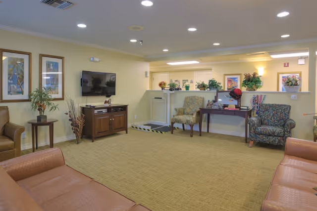 A spacious living room area in an assisted living facility with beige walls and carpet. The room features multiple upholstered chairs and sofas arranged around the space. A flat-screen TV is mounted on the wall above a wooden cabinet. There are framed pictures and plants decorating the room, and a small desk with a computer and decorative items is positioned against a half wall. The lighting is provided by recessed ceiling lights and wall sconces.