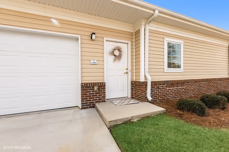 Exterior view of a beige and brick building showing a white door with a decorative wreath, a small concrete porch with a doormat, a window, and a closed white garage door. There are small bushes and green grass near the porch.