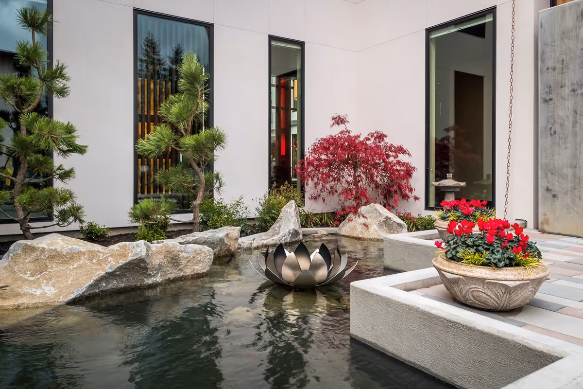 A tranquil outdoor courtyard with a small pond featuring a lotus-shaped metal sculpture in the center, surrounded by rocks, green pine trees, and a red-leaved shrub. There are two stone planters with red flowers on a tiled patio area next to the pond, and large windows of a building are visible in the background.