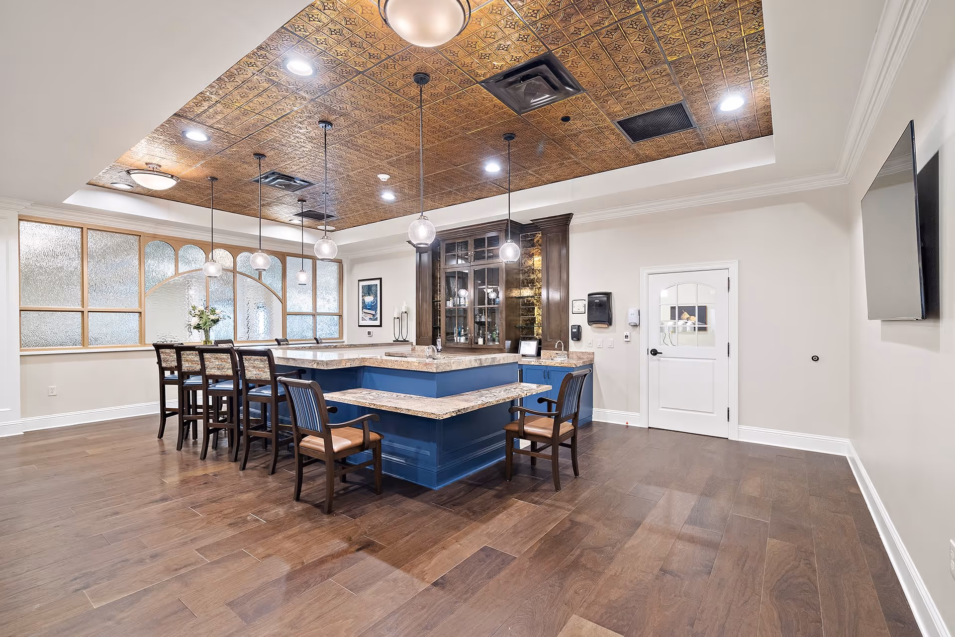 A spacious and elegant common area with a large blue island counter topped with marble, surrounded by wooden chairs with leather seats. The ceiling features decorative gold panels with recessed lighting and hanging globe pendant lights. The room has wooden flooring, a white door, a wall-mounted TV, and frosted glass windows with wooden frames.