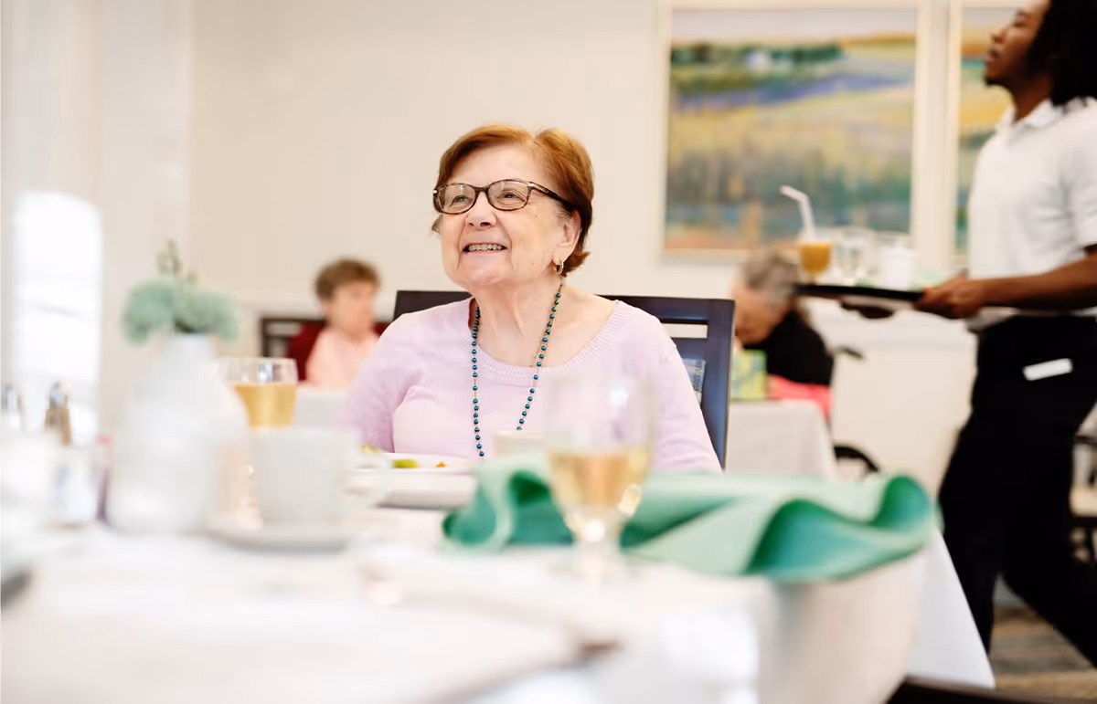 An elderly woman wearing glasses and a pink sweater is smiling while sitting at a dining table in a senior living facility. The table is set with a white tablecloth, a glass of water, and a green napkin. In the background, other elderly residents and a staff member carrying a tray are visible.