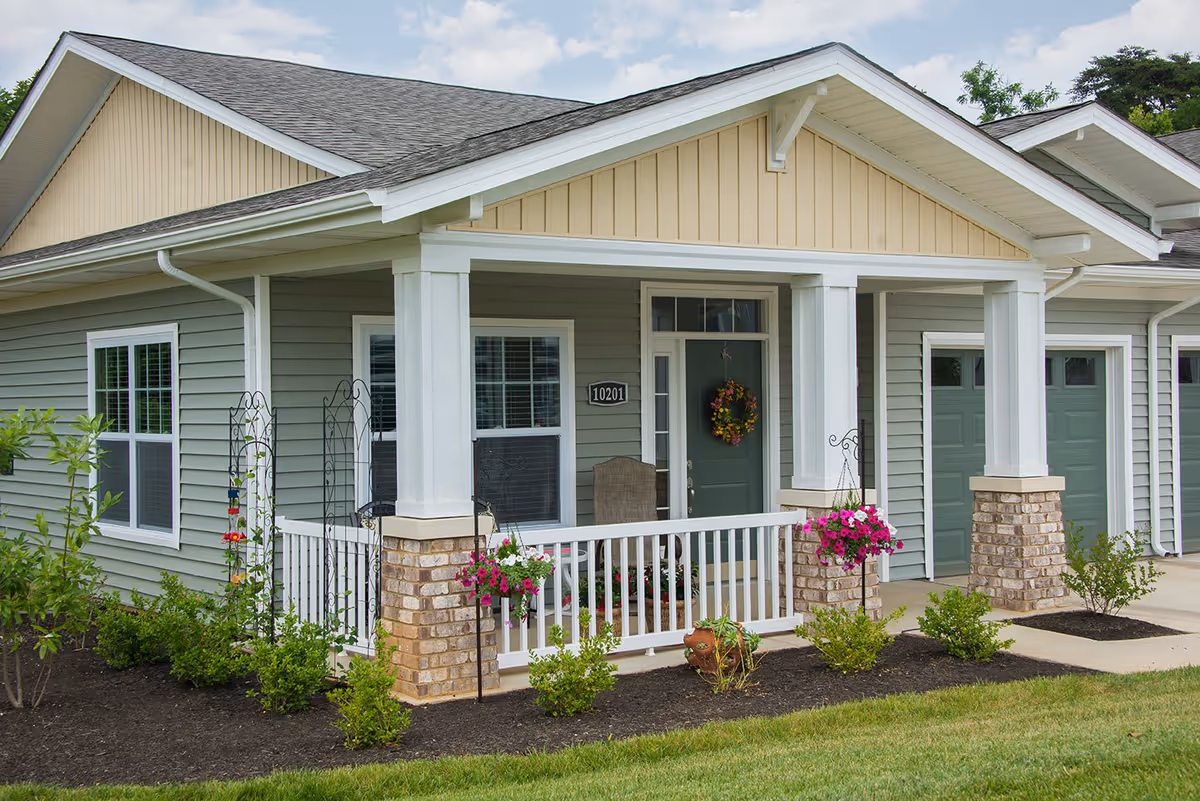 Exterior view of a single-story residential building with a covered porch featuring white railings and brick pillars. The porch has hanging flower baskets and a green front door decorated with a floral wreath. There are two windows with white frames and a garage door visible on the right side. The surrounding area includes neatly maintained landscaping with green shrubs and a lawn.