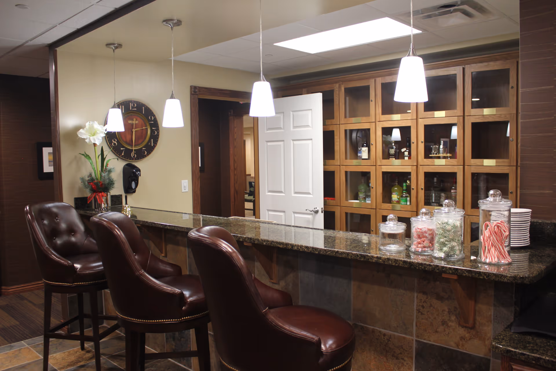 Interior view of a bar area with three brown leather bar stools, a granite countertop, and glass jars filled with candy canes and other sweets. Behind the bar is a wooden cabinet with glass doors containing bottles. A wall clock and a flower vase are visible on the left wall, and three pendant lights hang above the counter.