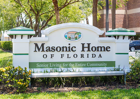 A large white sign with green accents reading 'Masonic Home of Florida' and 'Senior Living for the Entire Community' is displayed outdoors with trees and a building in the background.