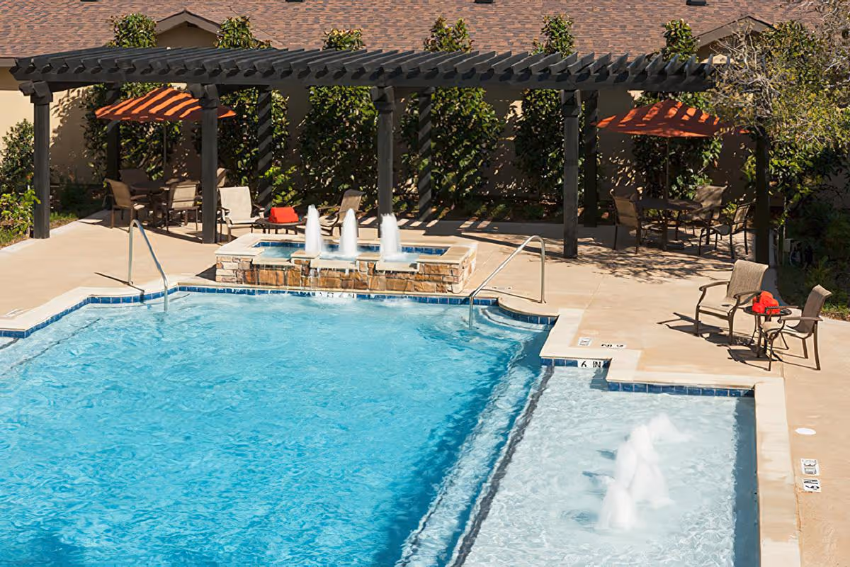 Outdoor swimming pool area with clear blue water, a smaller adjacent pool with water fountains, and a pergola with seating and tables shaded by orange umbrellas. The area is surrounded by greenery and a building with a shingled roof.