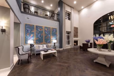 Spacious and elegant living room area with high ceilings, featuring a seating arrangement with a sofa, armchairs, a coffee table, and decorative wall art. There is a fireplace on the right side, a large window with curtains, and a round table with flower arrangements in the foreground. The room has a second-floor balcony with additional seating visible above.