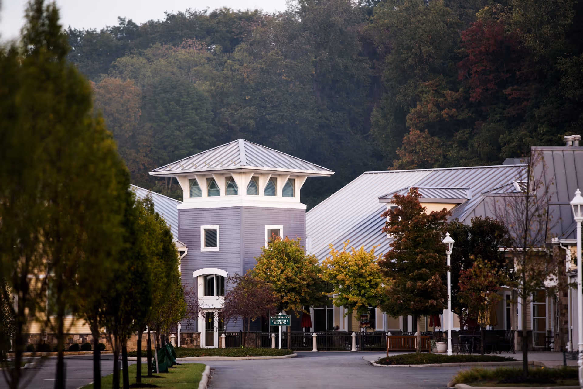Exterior view of Otterbein Granville SeniorLife Community showing a building with a distinctive tower and metal roofs, surrounded by trees with autumn foliage and a paved driveway.