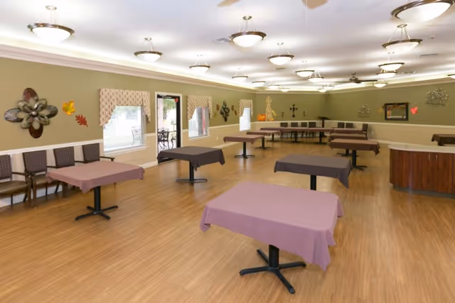 A spacious dining room with multiple tables covered in purple tablecloths and chairs arranged along the walls. The room has wooden flooring, light green walls decorated with metal wall art and seasonal decorations, and several ceiling lights providing bright illumination. Large windows and a door lead to an outdoor patio area.