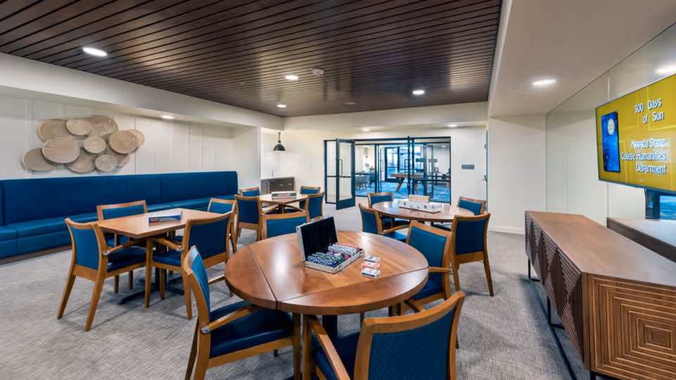 Bright communal activity/dining room with round and rectangular wooden tables, blue-upholstered chairs, a built-in bench, and a wall-mounted TV.