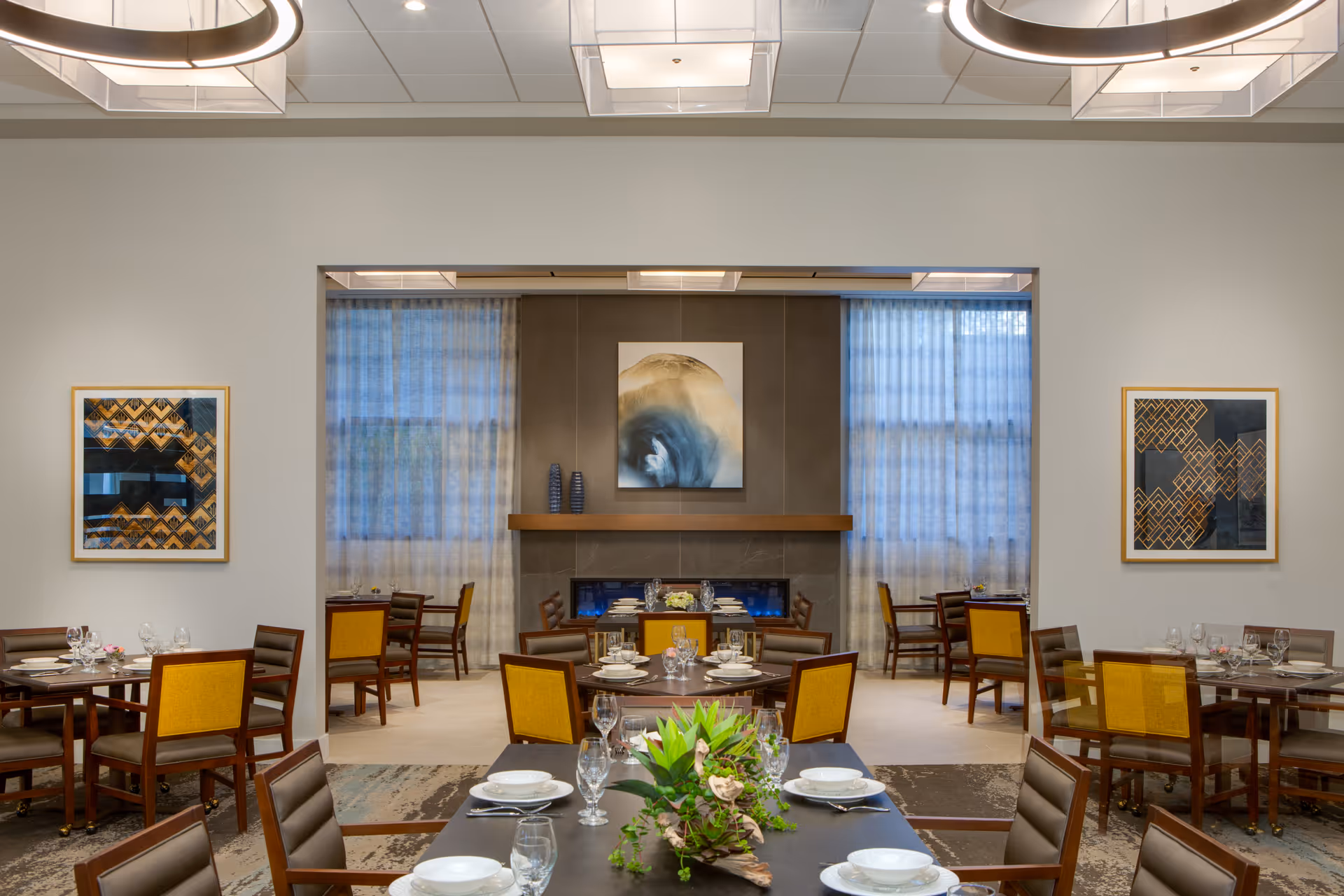 A modern dining room with multiple tables set with white plates, bowls, glasses, and silverware. The chairs have wooden frames with yellow and brown upholstery. The room features large windows with sheer curtains, abstract artwork on the walls, and a fireplace with a decorative mantle and vases. A green plant centerpiece is on the closest table.