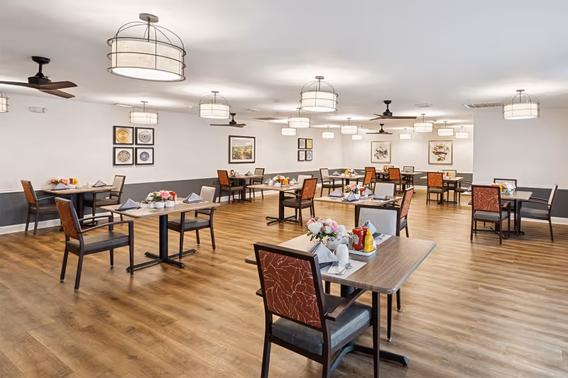A spacious dining room with multiple tables and chairs arranged neatly. Each table is set with napkins, condiments, and floral centerpieces. The room has wooden flooring, white walls with framed artwork, and ceiling lights with fabric shades. Ceiling fans are also visible.