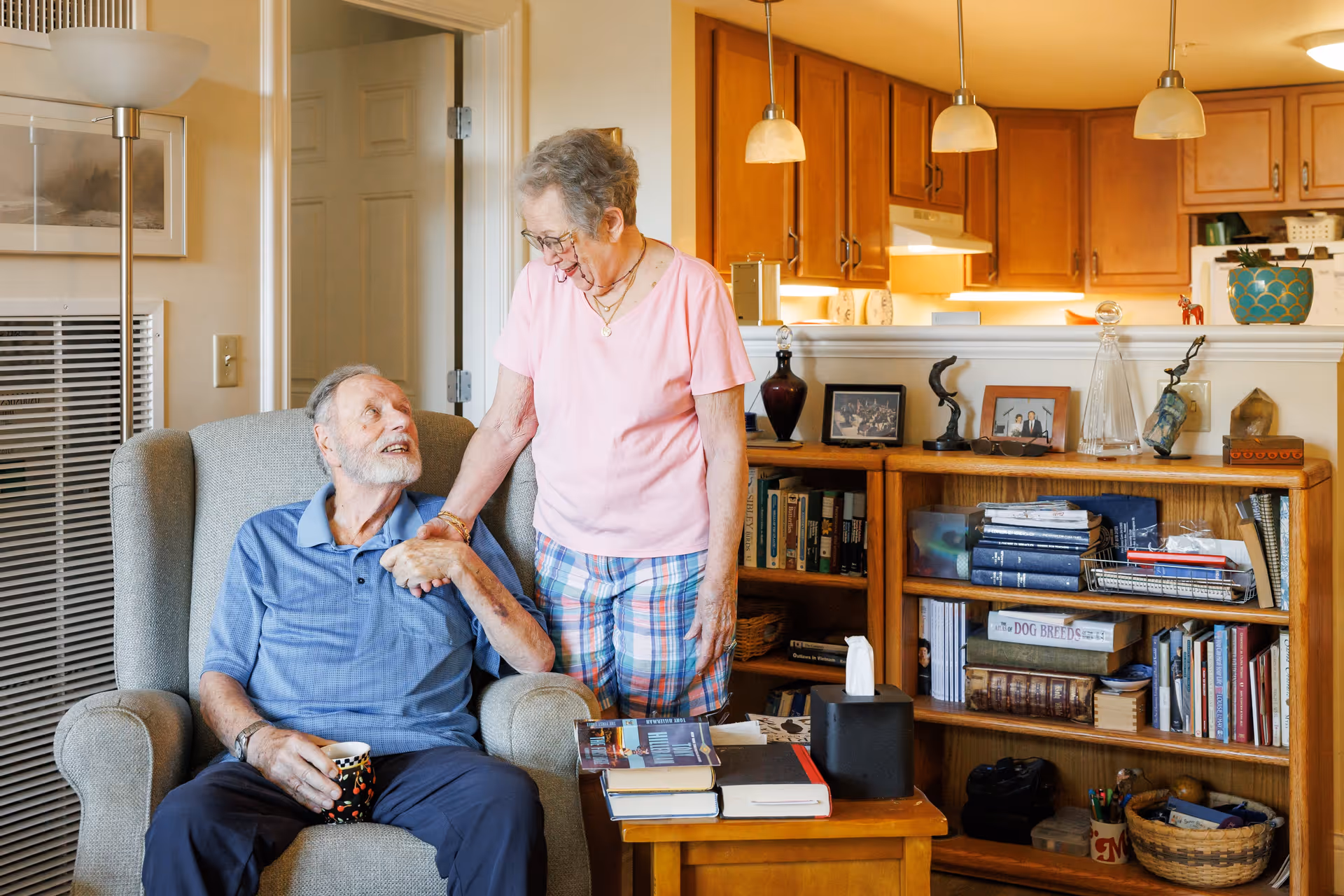 An elderly man sitting in a comfortable armchair holding a coffee mug, looking up and smiling at an elderly woman standing beside him holding his hand in a warm, cozy living room with wooden bookshelves filled with books and decorative items, and a kitchen area visible in the background.