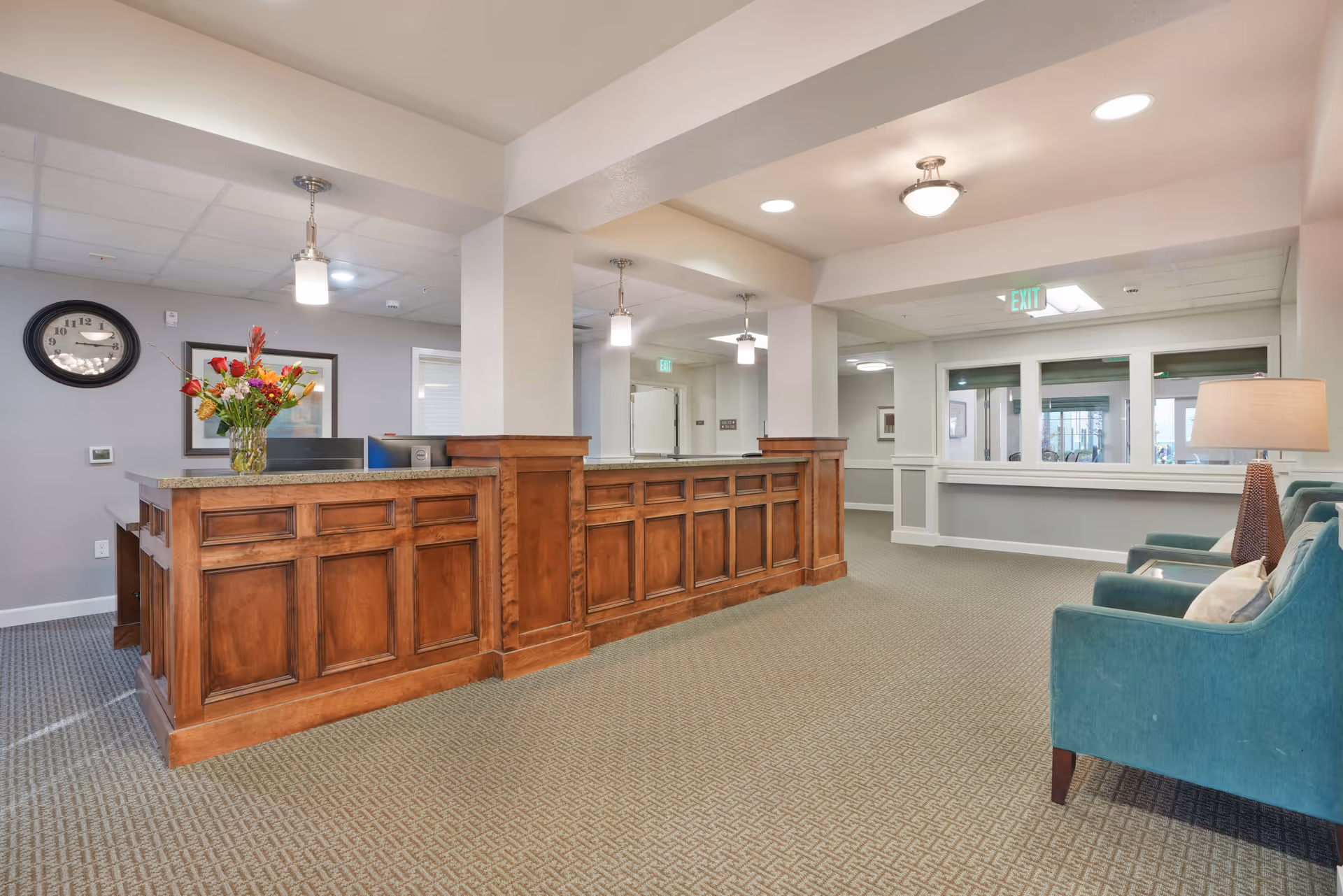 Reception area with a wooden front desk, a vase of flowers on the counter, a wall clock, and teal armchairs with a lamp in a well-lit room with carpeted floor.