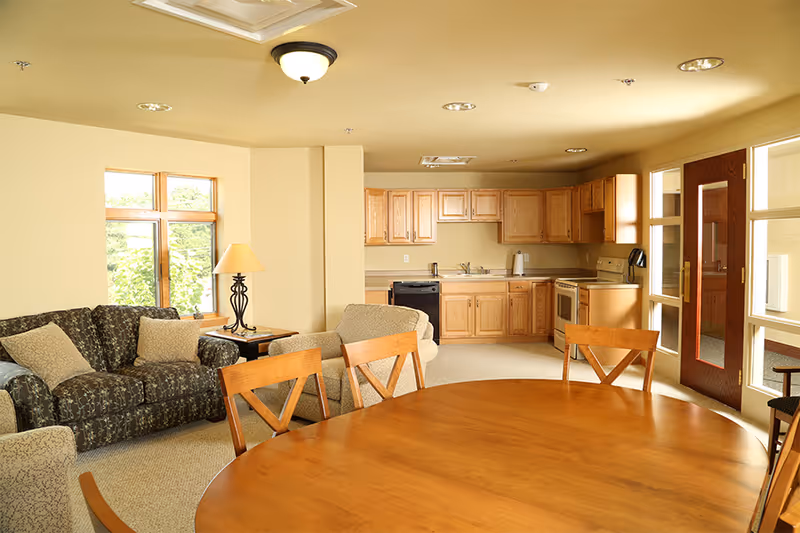 A bright and cozy living area with a round wooden dining table and four chairs in the foreground. Behind the table is a sitting area with a patterned sofa, an armchair, and a side table with a lamp. The background features a kitchen with wooden cabinets, a stove, dishwasher, and a sink. Large windows and a glass door allow natural light to fill the room.