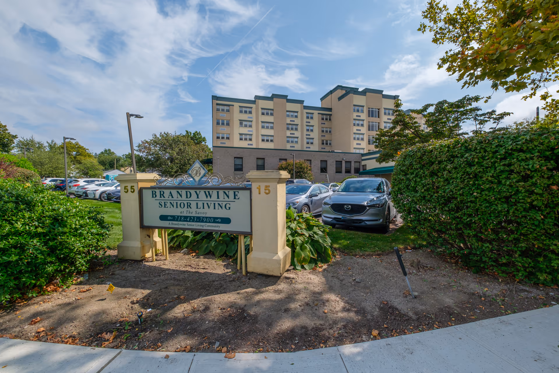 Exterior view of Brandywine Senior Living at The Savoy building with a sign in the foreground displaying the facility name and phone number. Several cars are parked near the building, and there are trees and bushes around the area under a partly cloudy sky.