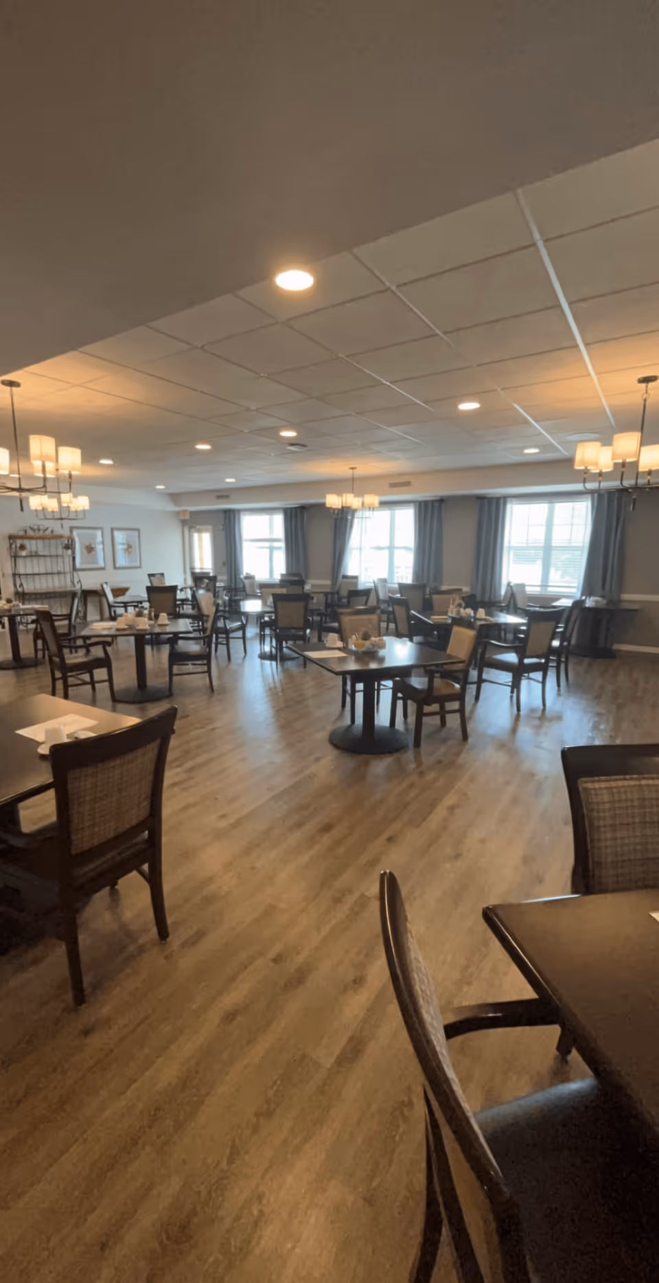 Spacious dining room with multiple tables and chairs arranged neatly on a wooden floor. The room is well-lit with ceiling lights and chandeliers, and large windows with curtains allow natural light to enter. The walls are decorated with framed artwork and there is a shelving unit in the background.