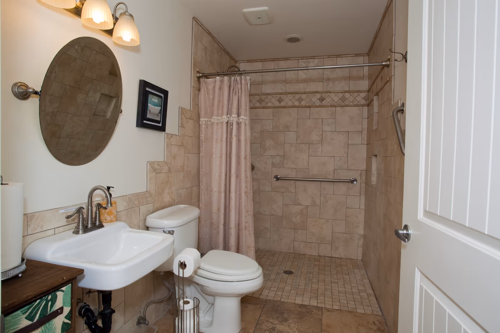 A bathroom with beige tiled walls and floor. It features a white toilet, a white sink with a silver faucet, an oval mirror above the sink, and a shower area with a pink patterned shower curtain and grab bars. There is a wooden cabinet with a green patterned drawer next to the sink and a roll of paper towels on top.