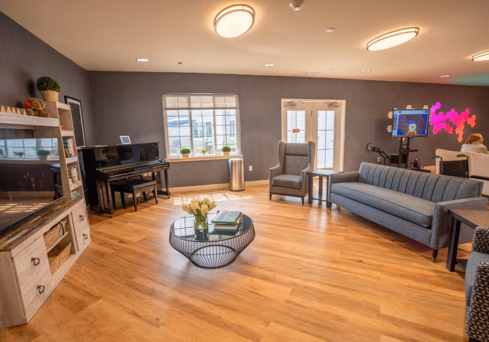 A bright and spacious living room with wooden flooring, a gray sofa, a patterned armchair, a round glass coffee table with books and flowers, a black upright piano, and a large window letting in natural light. There is a TV mounted on a wooden stand and a colorful light installation on the wall. A person is seated in the background near a screen.