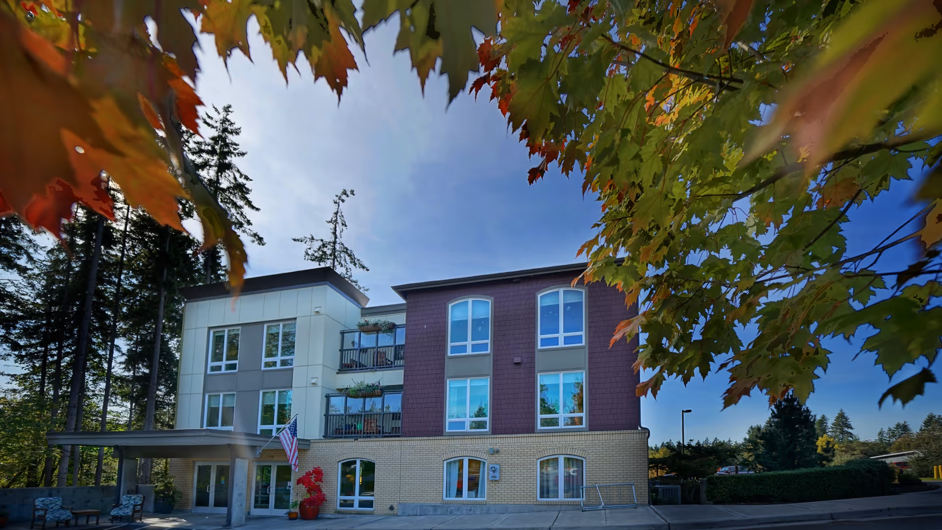 Exterior view of Bainbridge Senior Living Madrona House, a three-story building with large windows and balconies. The building is surrounded by trees with autumn-colored leaves in the foreground and a clear blue sky above.