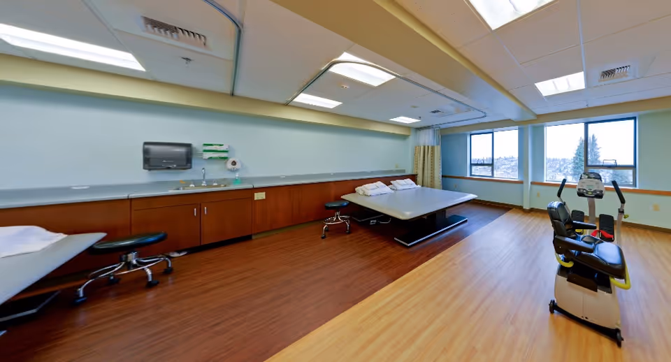 Bright rehabilitation room with treatment tables, rolling stools, cabinetry and sink along one wall and exercise equipment by the windows.