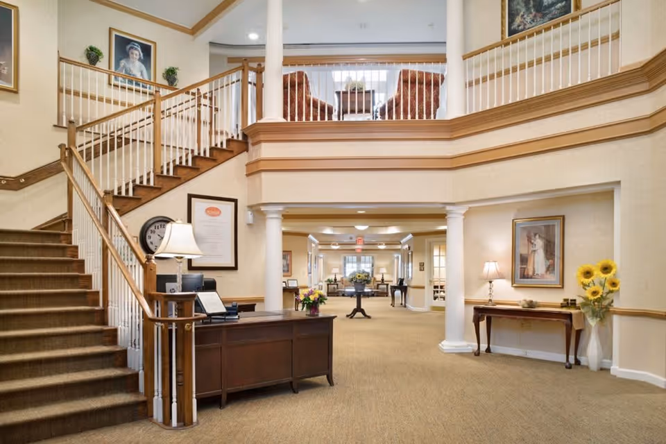 Interior view of a senior living facility lobby with a staircase on the left, a wooden reception desk with a lamp and flowers, and a hallway leading to a seating area. The space features beige walls, carpeted floors, framed artwork, and decorative columns.
