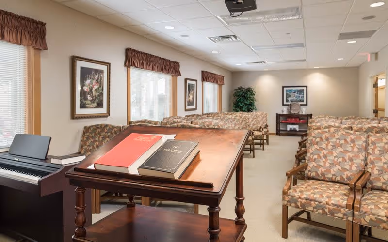 A well-lit room with rows of patterned armchairs arranged facing a wooden podium holding two large books. To the left, there is an electronic keyboard near windows with brown valances. The walls are decorated with framed floral artwork and a framed picture is visible on the far wall above a small table with a decorative plate and red books. A potted plant is in the corner.