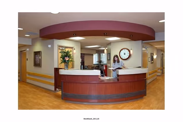 Reception area inside a senior living facility with a curved wooden front desk, a woman standing behind the desk looking at a book, a large clock on the wall, and a vase with flowers on the counter. The hallway extends to the right and left with beige walls and wooden handrails.